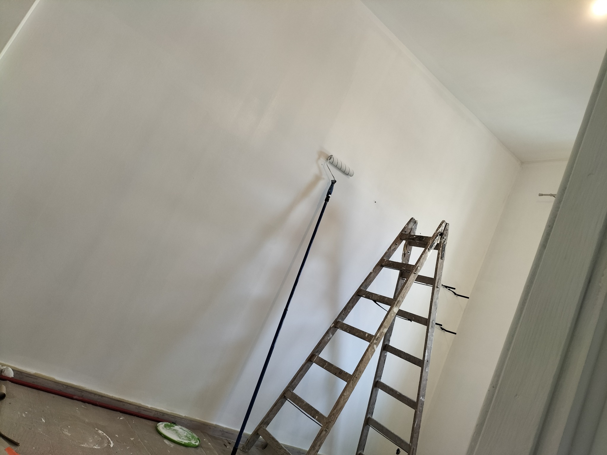 Świeżo pomalowana, biała ściana w pokoju, z widocznym wałkiem malarskim na długim kiju opartym o ścianę oraz drewnianą drabiną przystawną. Na podłodze widoczne ślady farby i plastikowa tacka.
