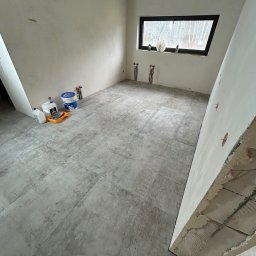 BudMex - Pokój po szpachlowaniu i wylewce, przygotowany do malowania. Widoczne narzędzia malarskie, okno i instalacje. Szare płytki podłogowe imitujące beton.