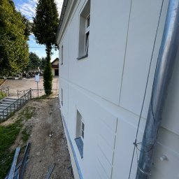 BudMex - Jasna elewacja budynku z dekoracyjnymi panelami, widok z boku, z rynną i elementami krajobrazu w tle. Świeżo pomalowana powierzchnia.