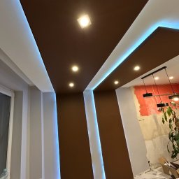 BudMex - Nowoczesny sufit podwieszany z brązowymi panelami i oświetleniem LED w wyremontowanym wnętrzu. Widoczne lampy i fragment ściany w trakcie prac.