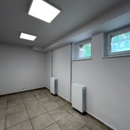 BudMex - Wnętrze po remoncie: szare płytki podłogowe, białe ściany z grzejnikami i oknami. Dwa kwadratowe panele LED na suficie. Minimalistyczny, odświeżony pokój.