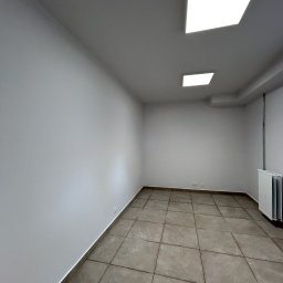 BudMex - Wnętrze po remoncie: gładkie, białe ściany, szara podłoga z płytek, nowoczesne oświetlenie sufitowe. Widoczny grzejnik. Minimalistyczny design.