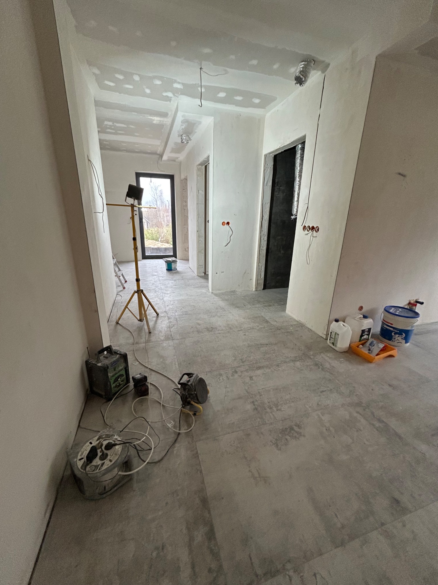 Wnętrze w trakcie prac wykończeniowych: surowe ściany z widocznymi puszkami elektrycznymi, gładzie na suficie, lampa budowlana i narzędzia na podłodze.