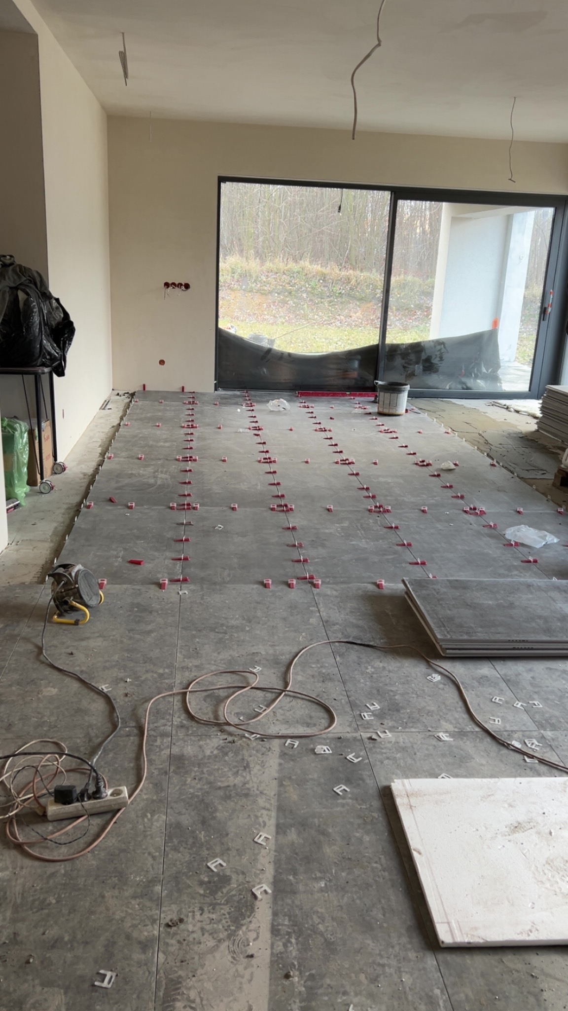 Szare płytki podłogowe w trakcie układania, z użyciem czerwonych krzyżyków dystansowych i białych klinów. Widoczne kable i narzędzia, duże okno w tle.