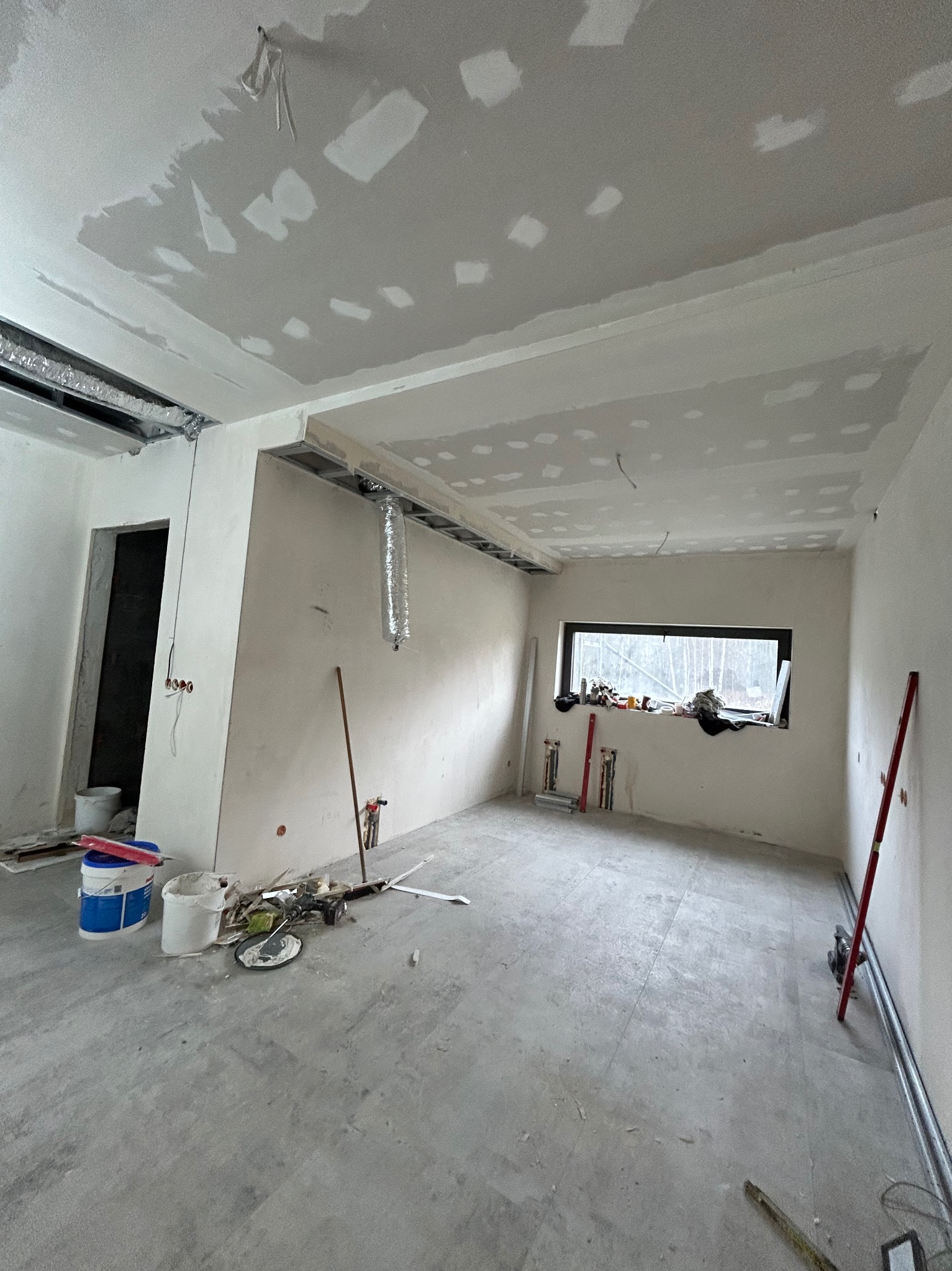 Wnętrze w trakcie prac wykończeniowych: sufit z płyt karton-gips, ściany po szpachlowaniu, widoczne narzędzia i materiały budowlane. Okno z widokiem na zewnątrz.