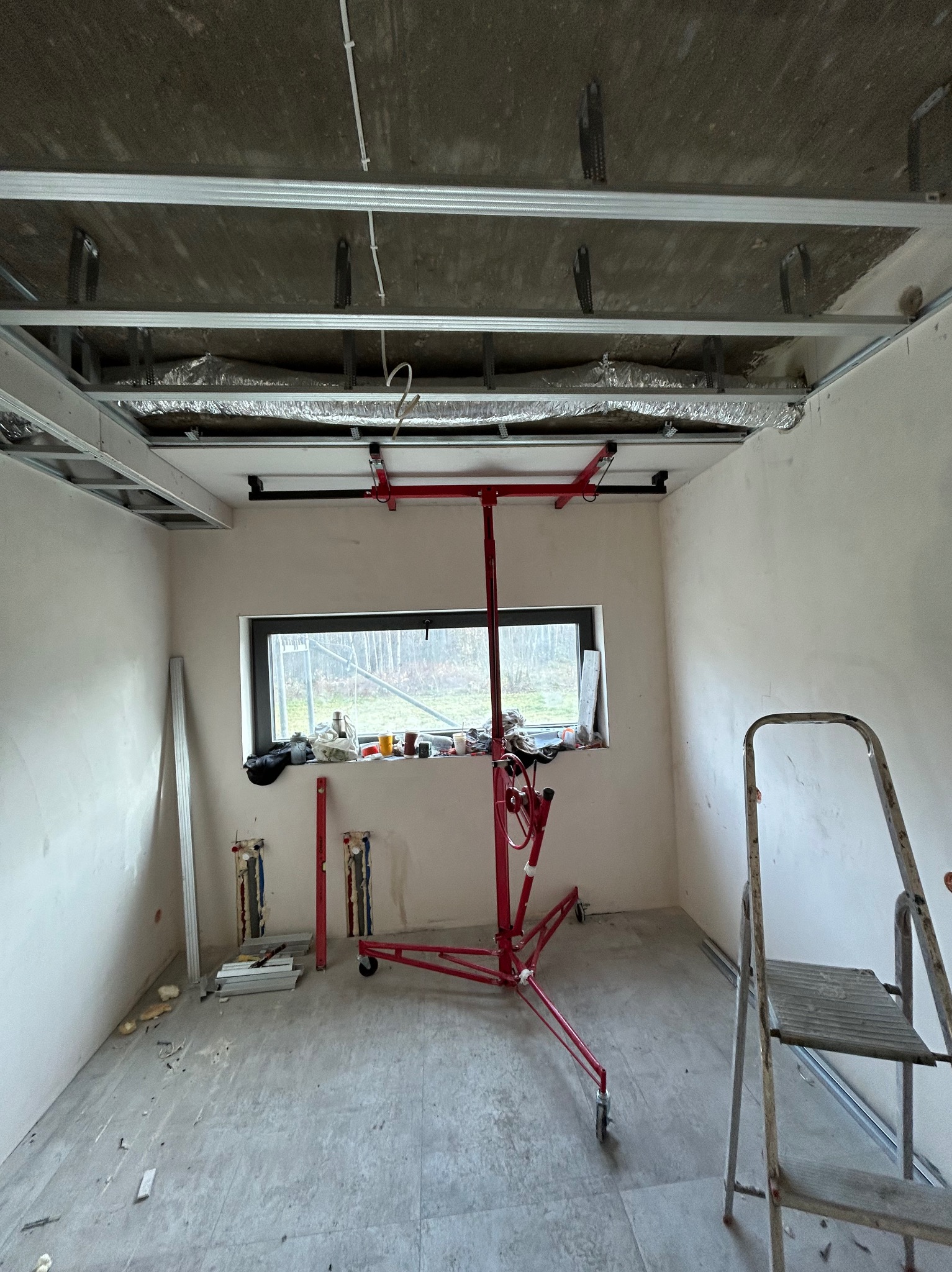 Montaż sufitu podwieszanego z płyt gipsowo-kartonowych. Widoczny stelaż, częściowo zamontowane płyty i podnośnik do płyt. W tle okno z widokiem na zewnątrz.