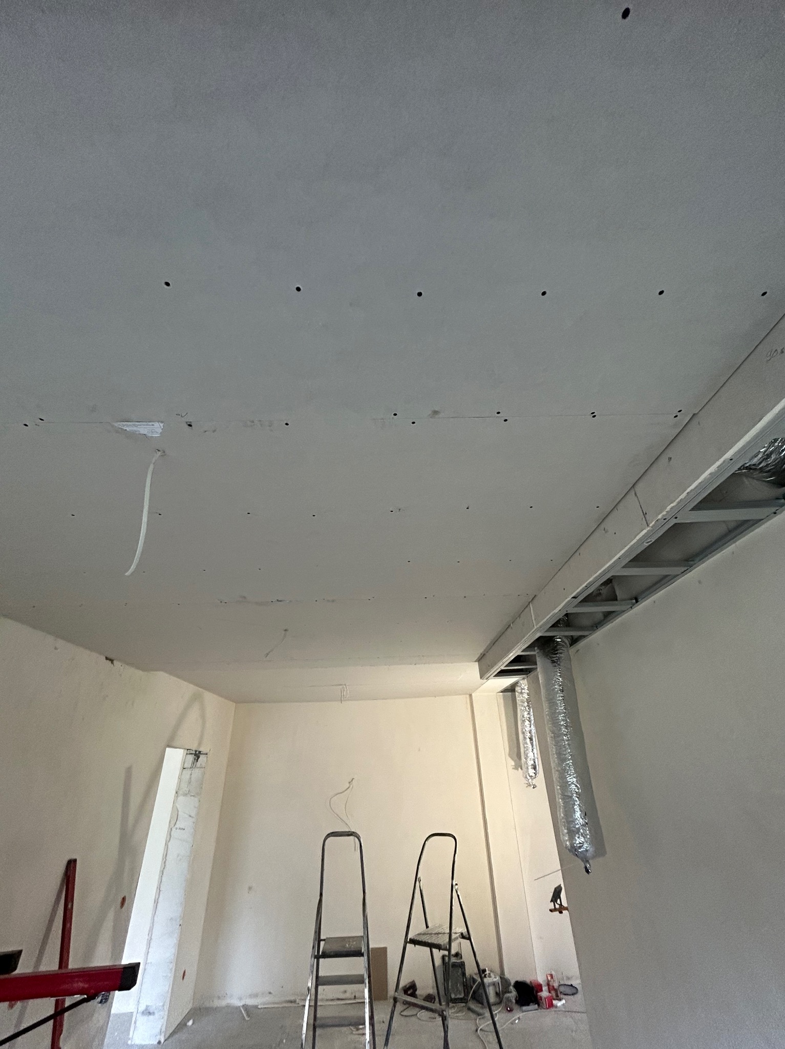 Montaż sufitu podwieszanego z płyt karton-gips, widoczne profile i instalacja wentylacyjna. Dwie drabiny w pomieszczeniu, ściany przygotowane do malowania.