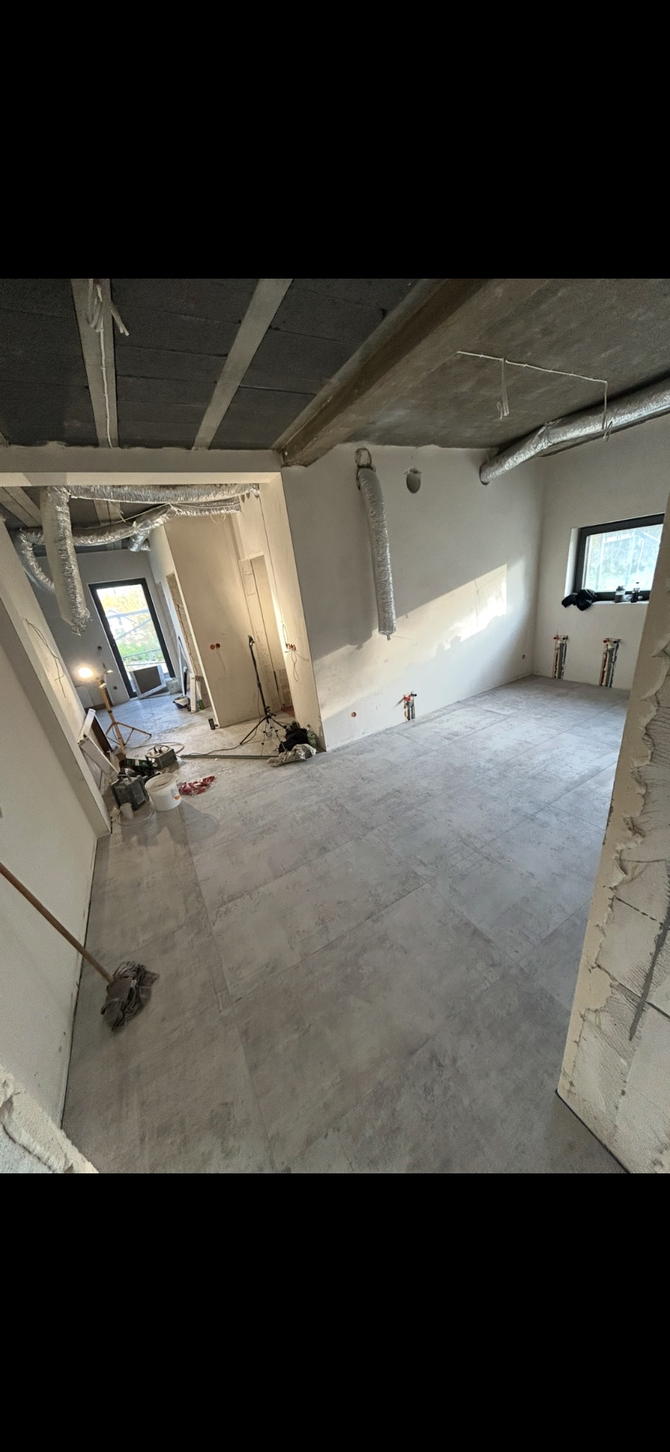 Wnętrze w trakcie prac wykończeniowych: szare płytki podłogowe, surowy betonowy sufit z widocznymi instalacjami wentylacyjnymi, ściany przygotowane do malowania.