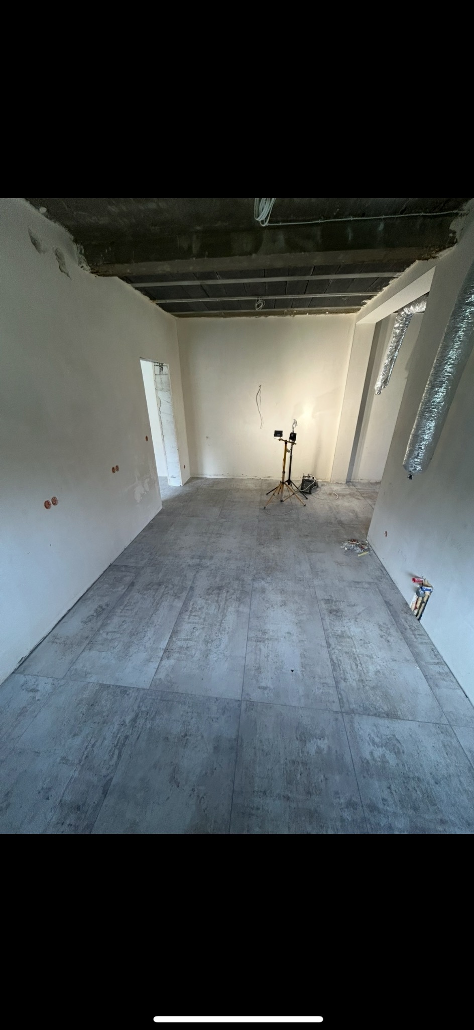 Wnętrze w trakcie remontu: szare płytki podłogowe, ściany po szpachlowaniu, widoczne instalacje. Na środku pomieszczenia statyw z lampą oświetlającą przestrzeń.