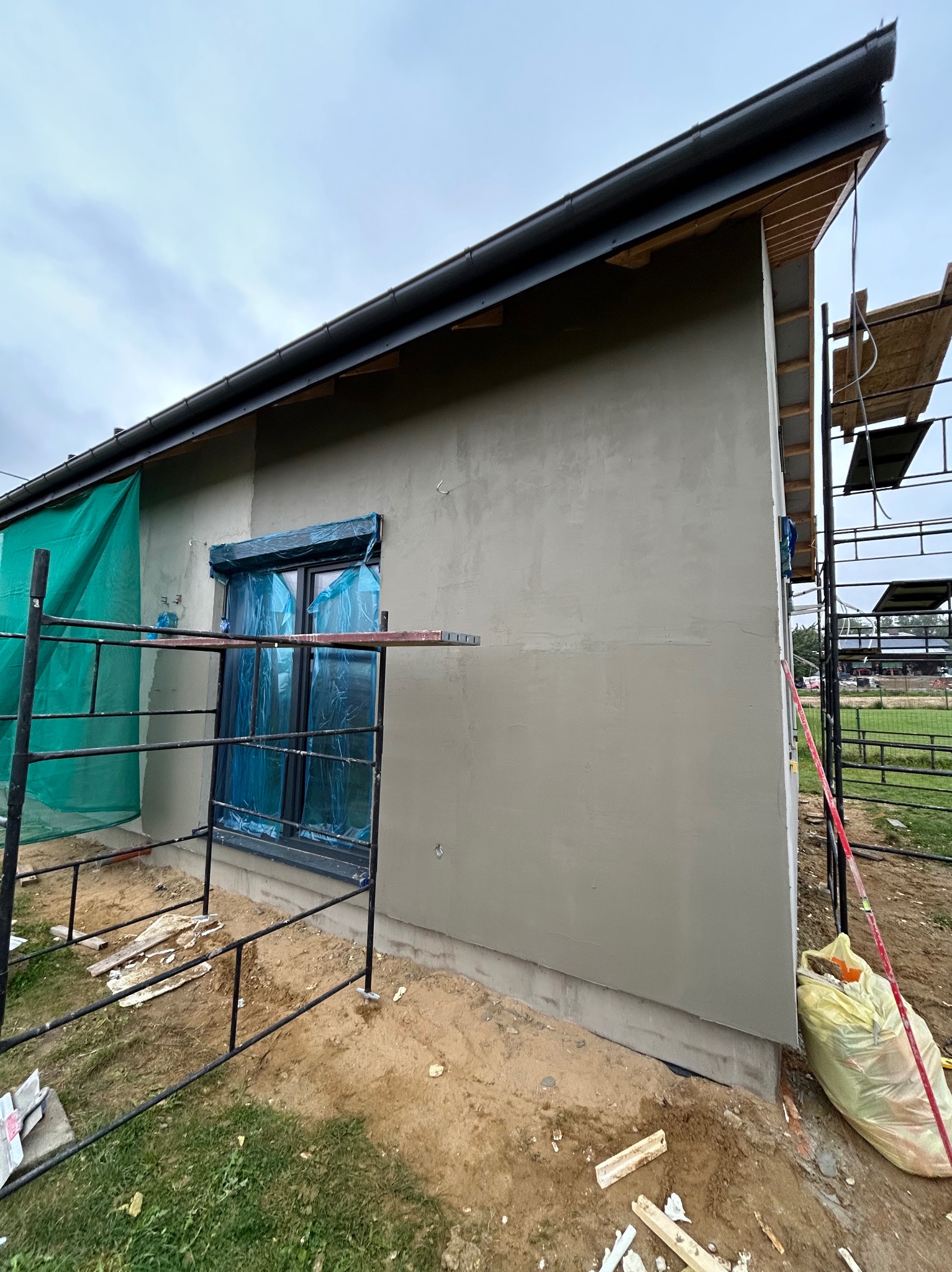 Ściana budynku po nałożeniu szarej gładzi, okno zabezpieczone folią, widoczne rusztowanie i teren budowy. Prace wykończeniowe elewacji w toku.