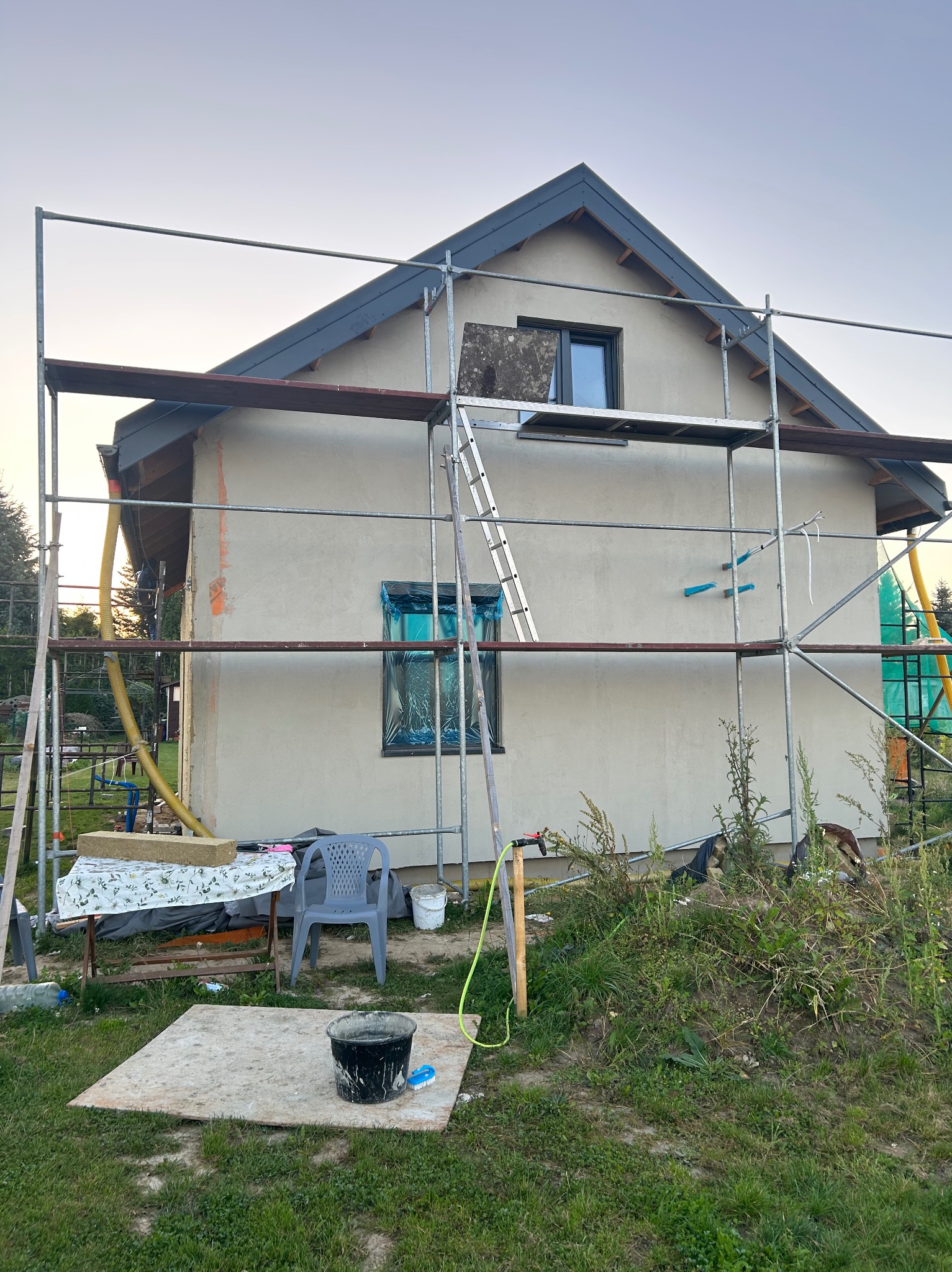 Dom jednorodzinny w trakcie prac elewacyjnych, otoczony rusztowaniem. Okna zabezpieczone folią, widoczne narzędzia i materiały budowlane. Prace w toku.