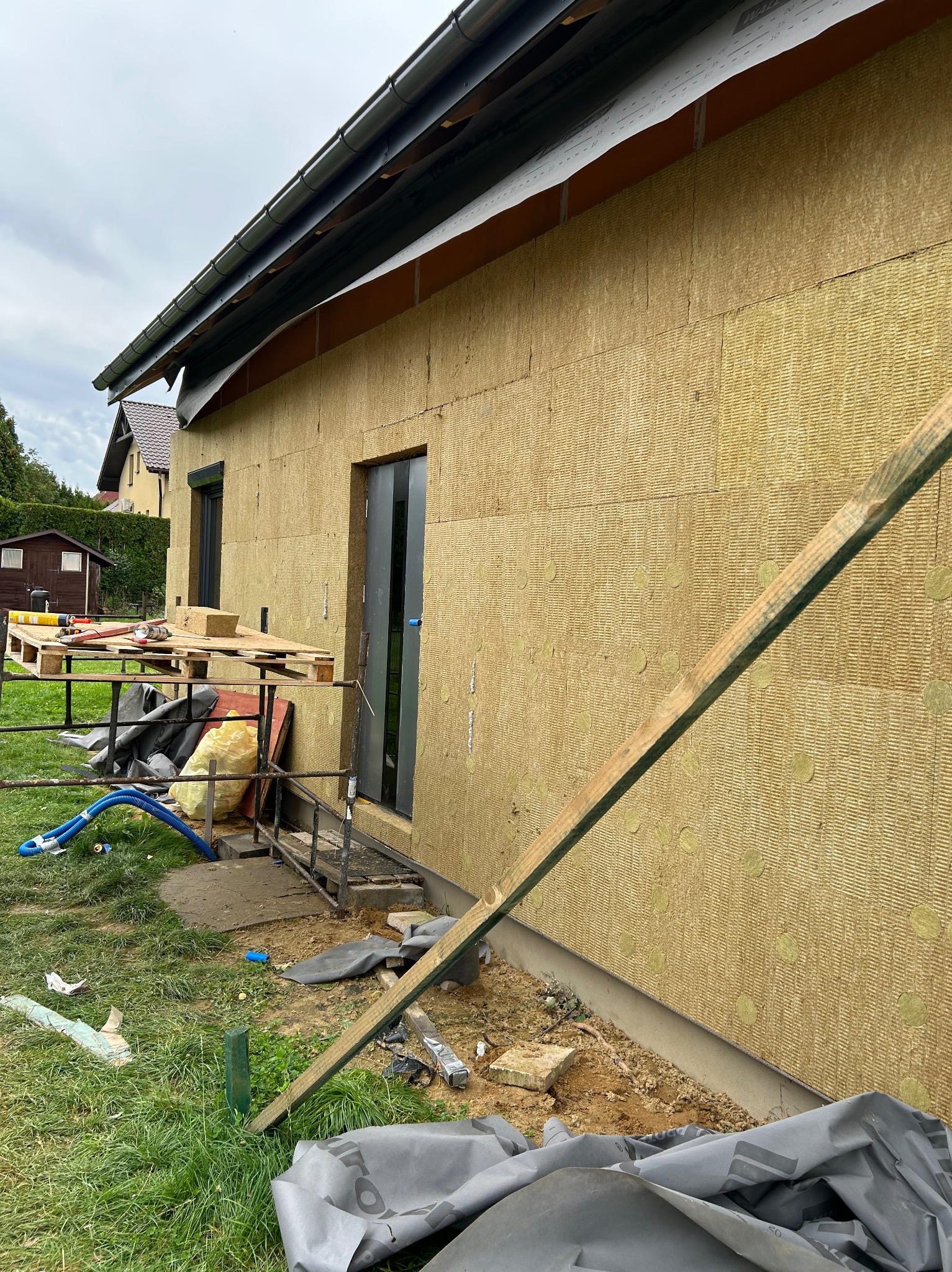 Ocieplona ściana budynku z widoczną wełną mineralną, rusztowaniem i narzędziami. Etap prac przed nałożeniem warstwy wykończeniowej elewacji. Widok z zewnątrz.