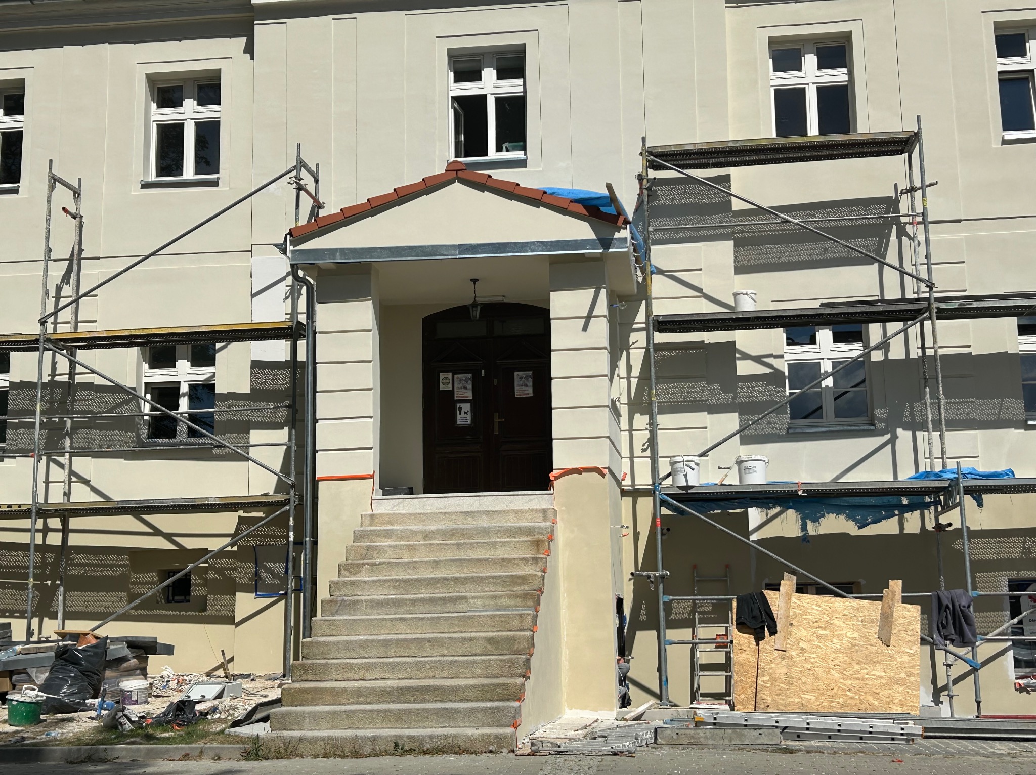 Budynek w trakcie prac elewacyjnych. Widoczne rusztowania, świeżo pomalowana fasada i schody wejściowe. Prace wykończeniowe w toku, z widocznymi narzędziami i materiałami.