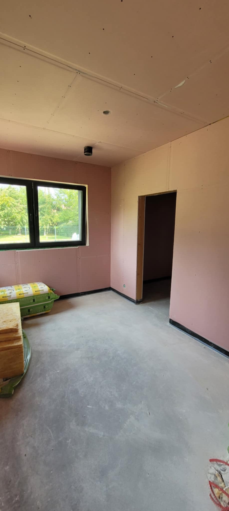 Wnętrze w trakcie wykańczania: ściany obłożone płytami karton-gips w odcieniach różu, widoczne materiały izolacyjne i surowa podłoga. Okno z widokiem na zieleń.