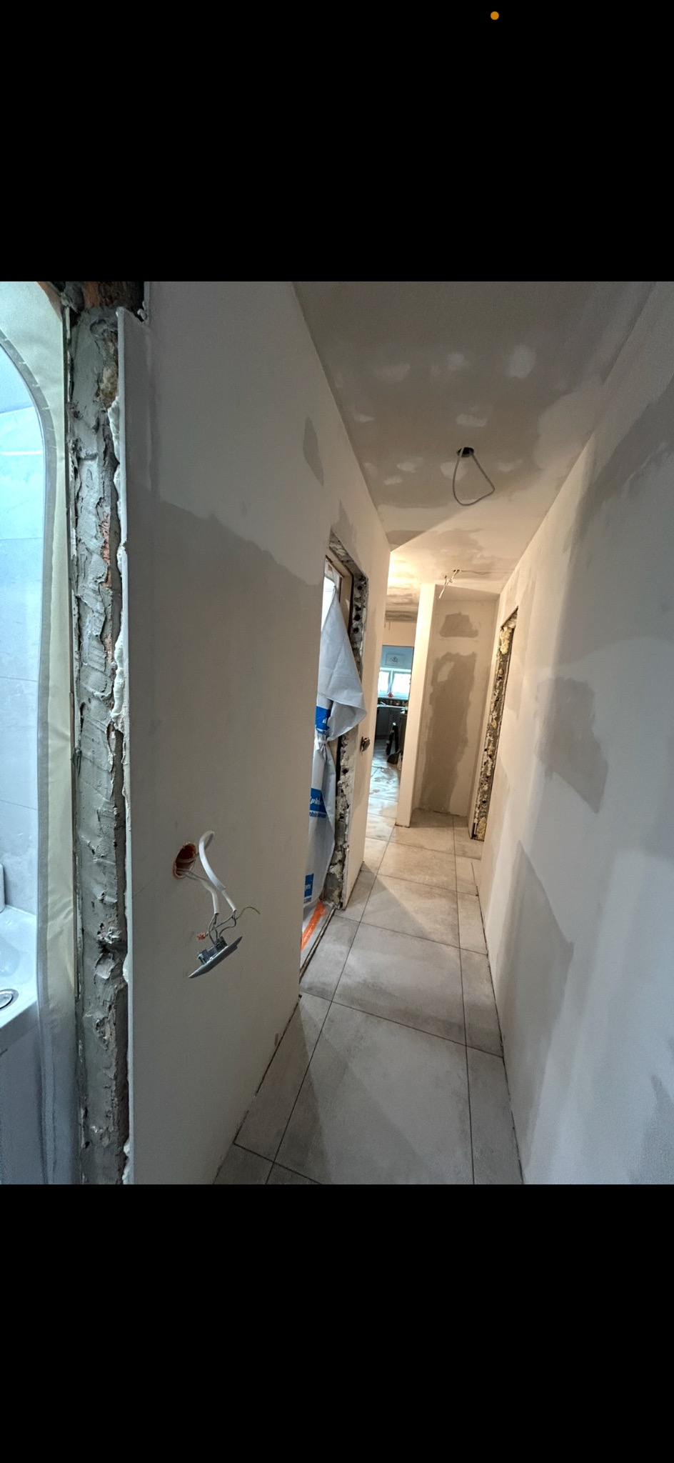 Korytarz w trakcie remontu: ściany po szpachlowaniu, widoczne przewody elektryczne, płytki na podłodze. Fragment łazienki z wanną po lewej stronie.