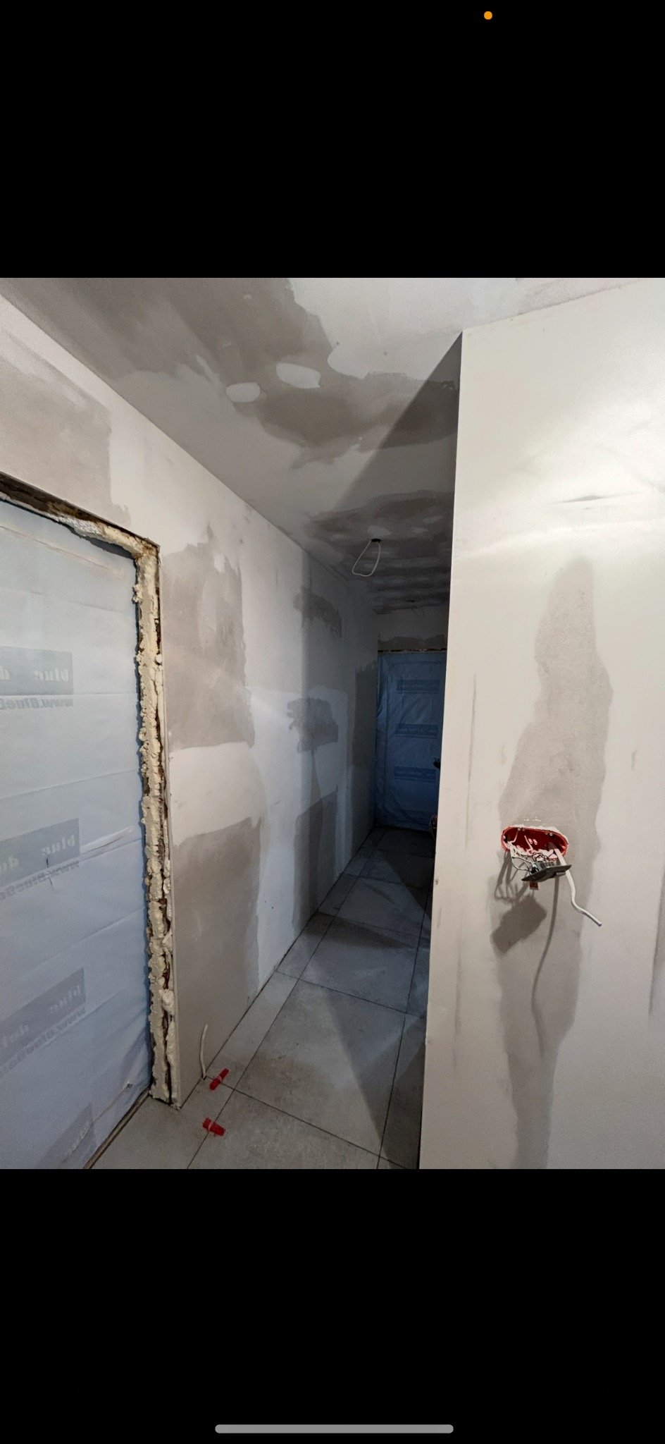 Korytarz w trakcie prac wykończeniowych: ściany po szpachlowaniu, widoczne puszki elektryczne, zabezpieczone drzwi. Podłoga wyłożona płytkami.