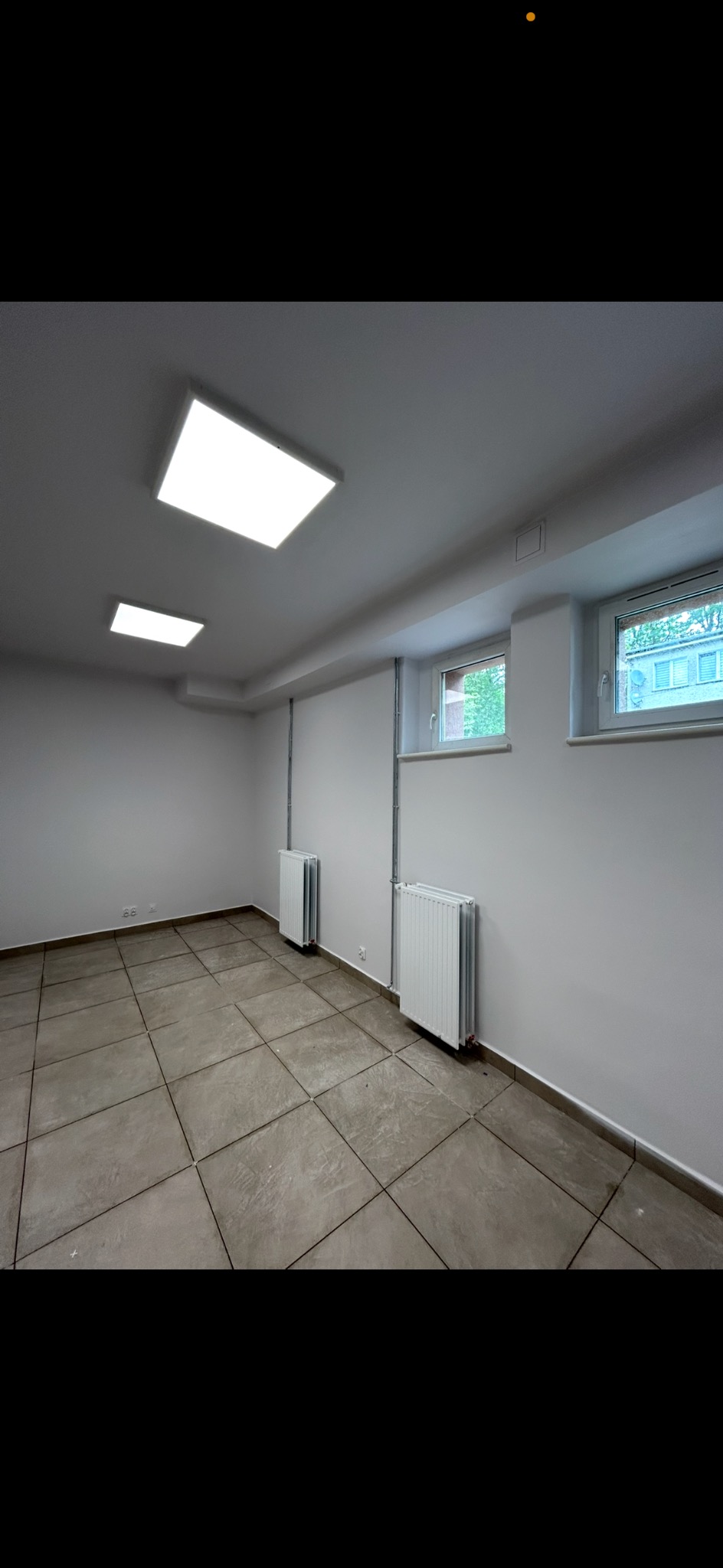 Wnętrze po remoncie: szare płytki podłogowe, białe ściany z grzejnikami i oknami. Dwa kwadratowe panele LED na suficie. Minimalistyczny, odświeżony pokój.