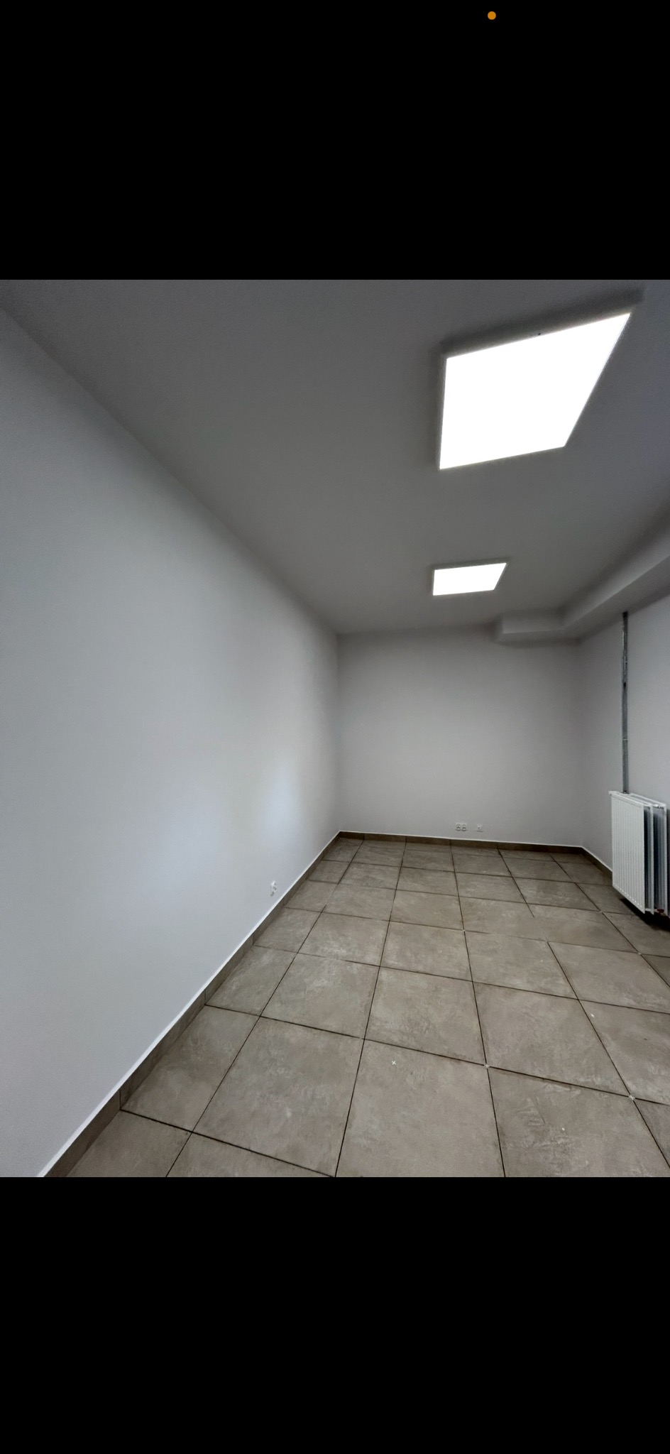 Wnętrze po remoncie: gładkie, białe ściany, szara podłoga z płytek, nowoczesne oświetlenie sufitowe. Widoczny grzejnik. Minimalistyczny design.