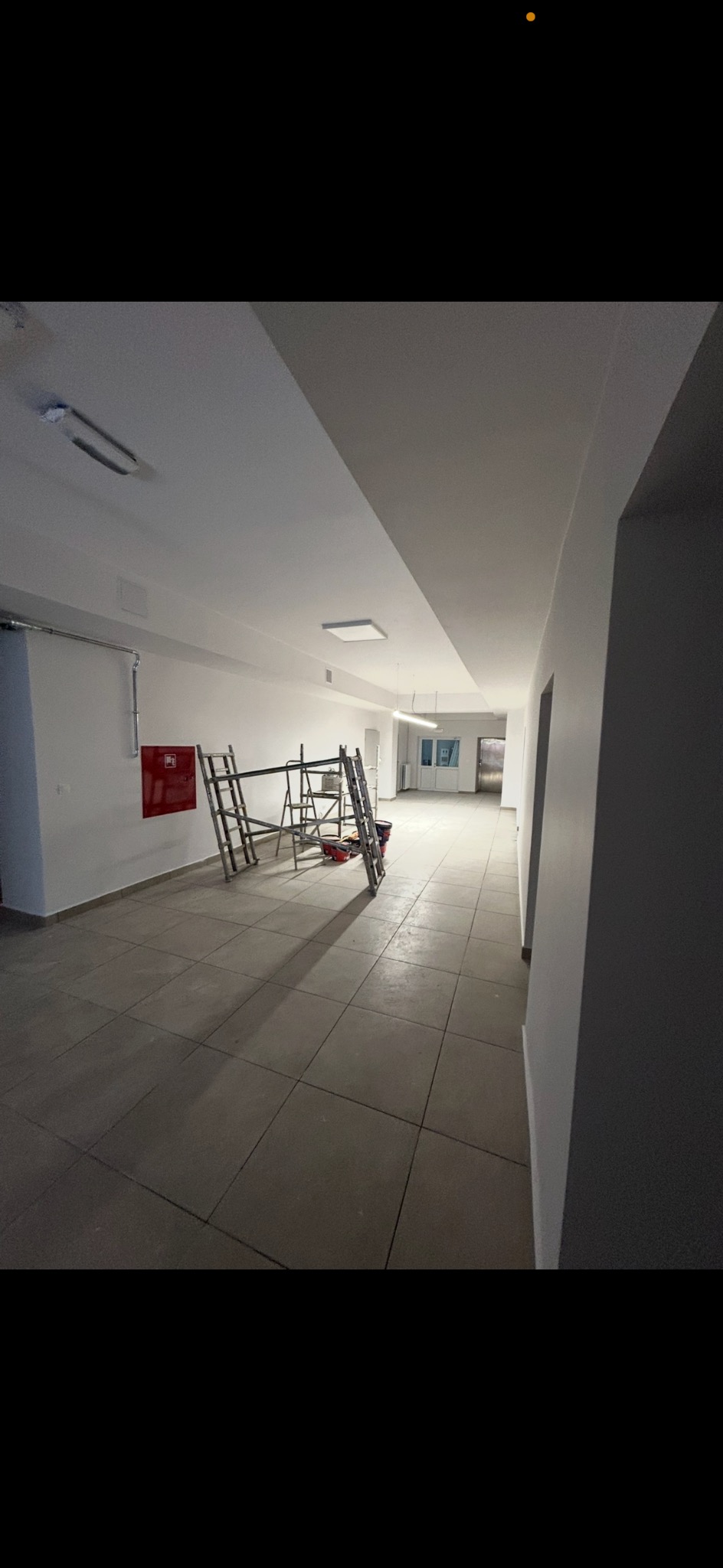 Wnętrze korytarza w trakcie prac wykończeniowych. Widoczne rusztowania, białe ściany i szara podłoga z płytek. Czerwona skrzynka hydrantowa na ścianie.