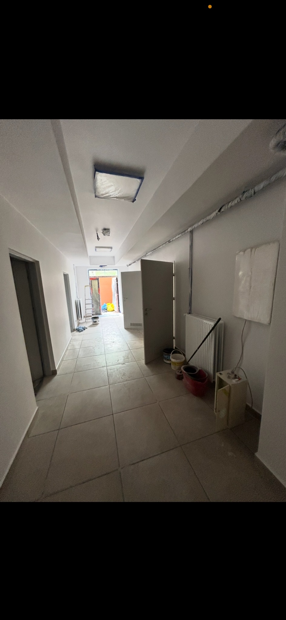 Korytarz w trakcie remontu: świeżo pomalowane ściany, zabezpieczone lampy, widoczne narzędzia malarskie i grzejnik. Jasne wnętrze z płytkami na podłodze.