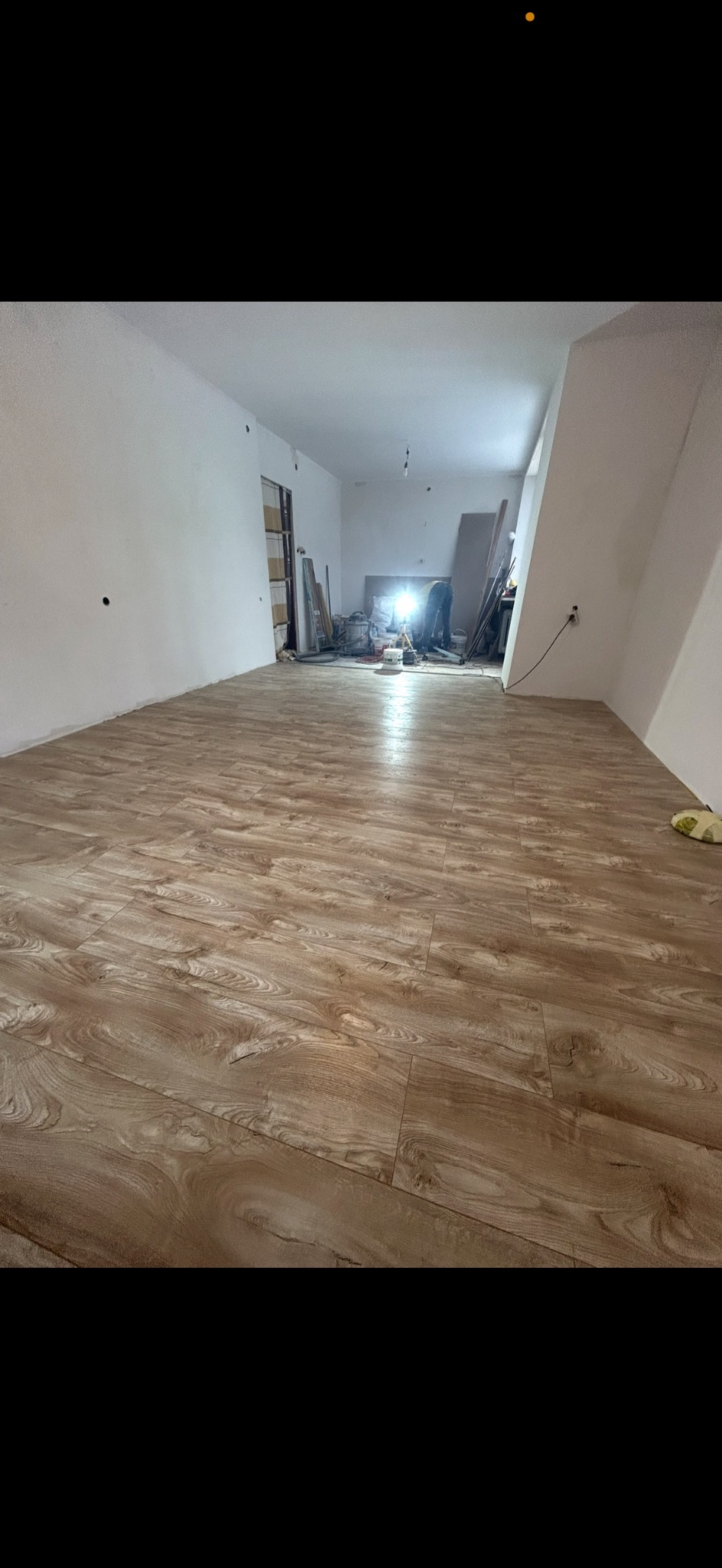 Jasne panele podłogowe ułożone w pustym pokoju z białymi ścianami. W tle widoczne narzędzia i materiały budowlane, sugerujące trwający remont.