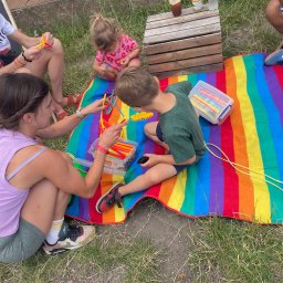 Bajkowooo.kreatywneanimacje Wiktoria Eliasz - Animator prowadzi zajęcia plastyczne na kocu w tęczowe pasy. Dzieci i dorosły tworzą konstrukcje z kolorowych patyczków. Na trawie drewniana skrzynka i napoje.