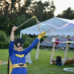 Bajkowooo.kreatywneanimacje Wiktoria Eliasz - Kobieta w kostiumie nietoperza tworzy ogromne bańki mydlane na tle zieleni i białego namiotu. Atrakcja dla dzieci na imprezie plenerowej.