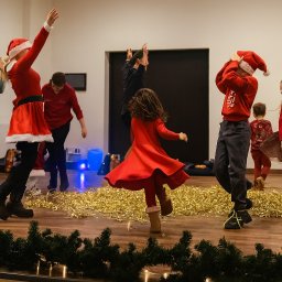 Bajkowooo.kreatywneanimacje Wiktoria Eliasz - Świąteczny występ dzieci na scenie: dziewczynka w stroju Elsy, dzieci w czerwonych strojach Mikołaja, dekoracje z lampek i girland. Radosna atmosfera.