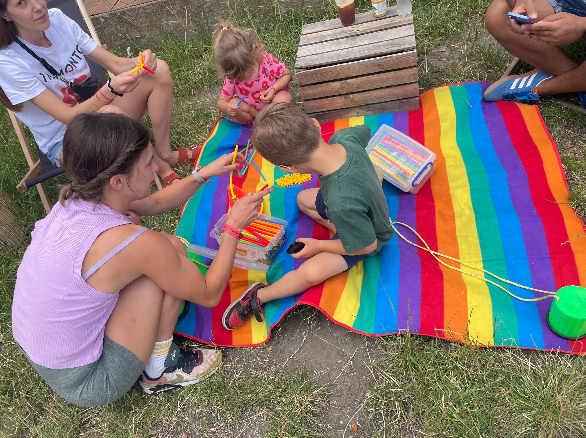 Animator prowadzi zajęcia plastyczne na kocu w tęczowe pasy. Dzieci i dorosły tworzą konstrukcje z kolorowych patyczków. Na trawie drewniana skrzynka i napoje.
