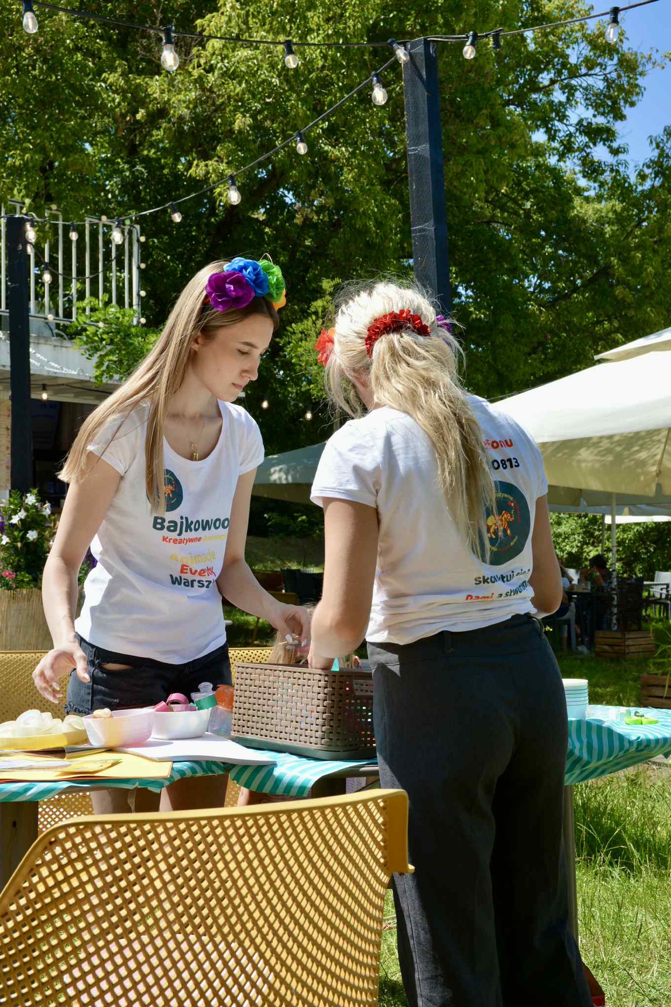 Dwie animatorki w koszulkach z logo firmy przygotowują materiały do warsztatów kreatywnych na zewnątrz. Widoczne kolorowe ozdoby do włosów i rekwizyty.
