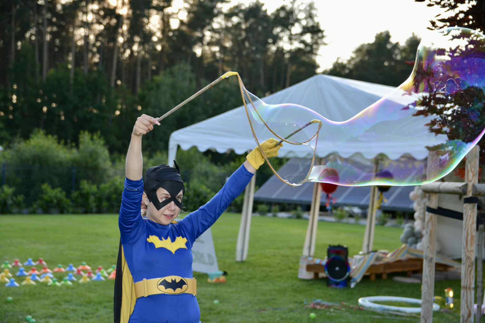 Kobieta w kostiumie nietoperza tworzy ogromne bańki mydlane na tle zieleni i białego namiotu. Atrakcja dla dzieci na imprezie plenerowej.