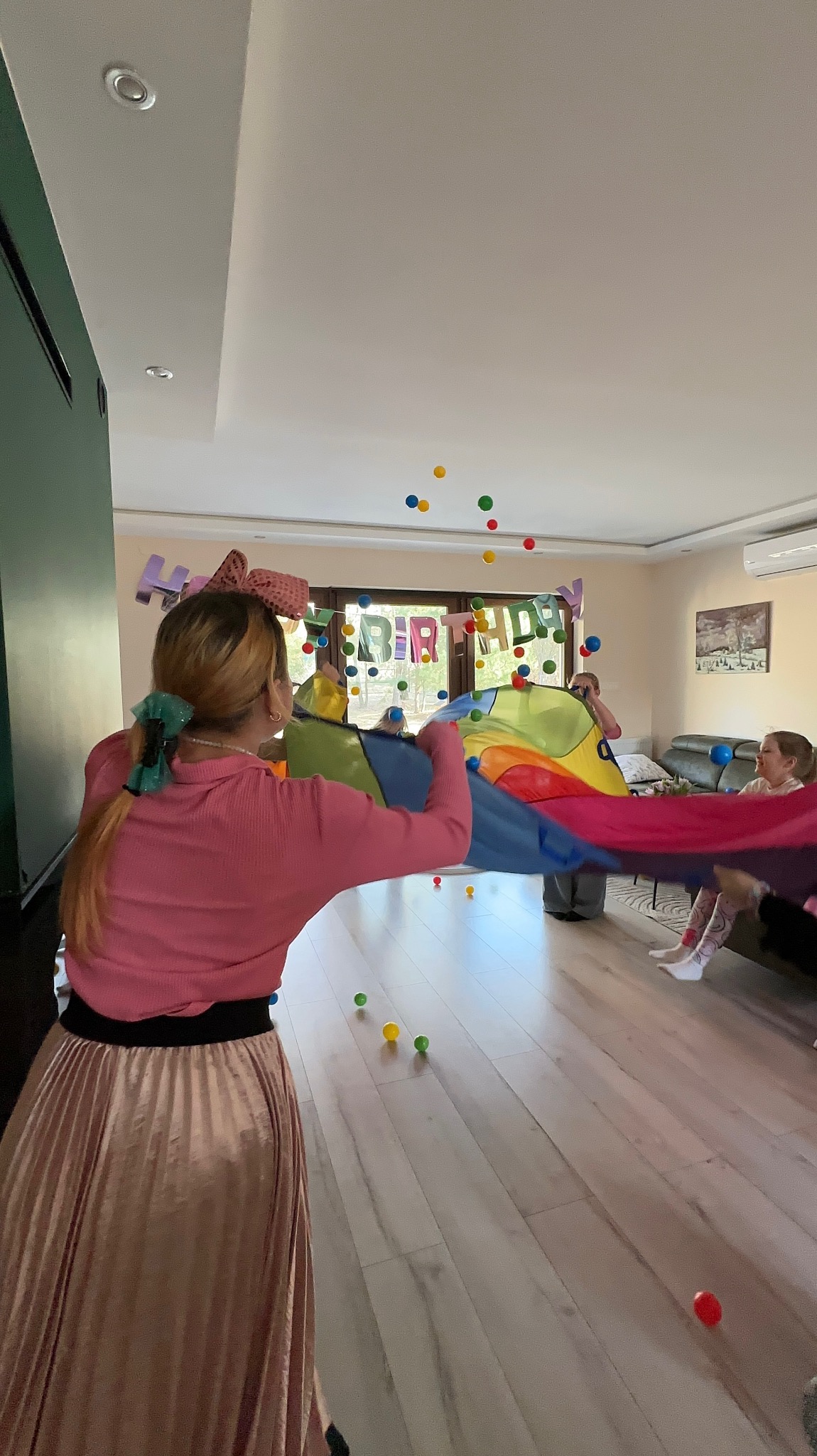 Animator z tęczową chustą animacyjną i unoszącymi się kolorowymi piłeczkami podczas urodzinowego przyjęcia w jasnym salonie.