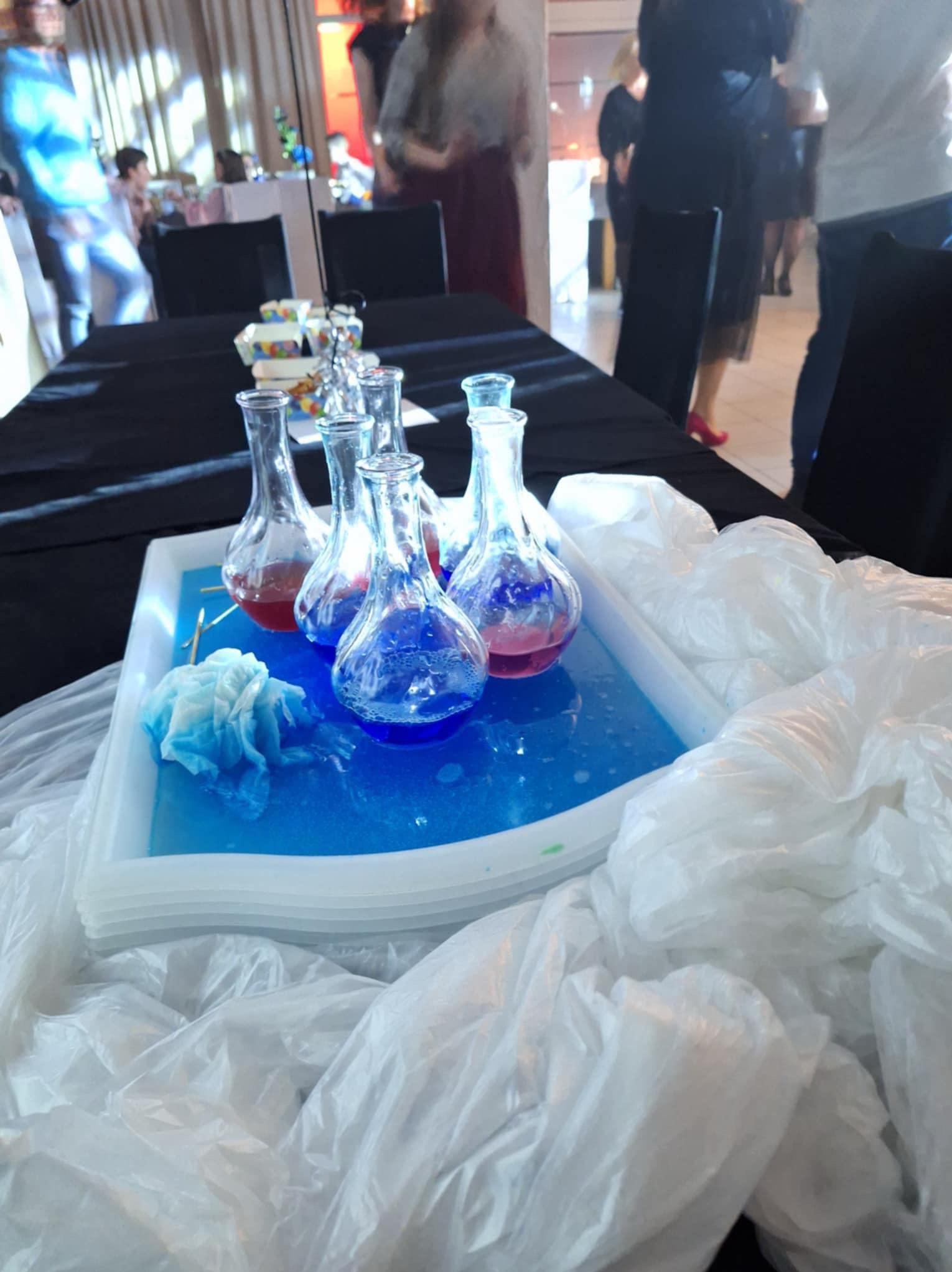 Eksperymenty chemiczne dla dzieci: probówki z kolorowymi płynami na tacy, otoczone dekoracjami z folii, w tle sala z ludźmi.