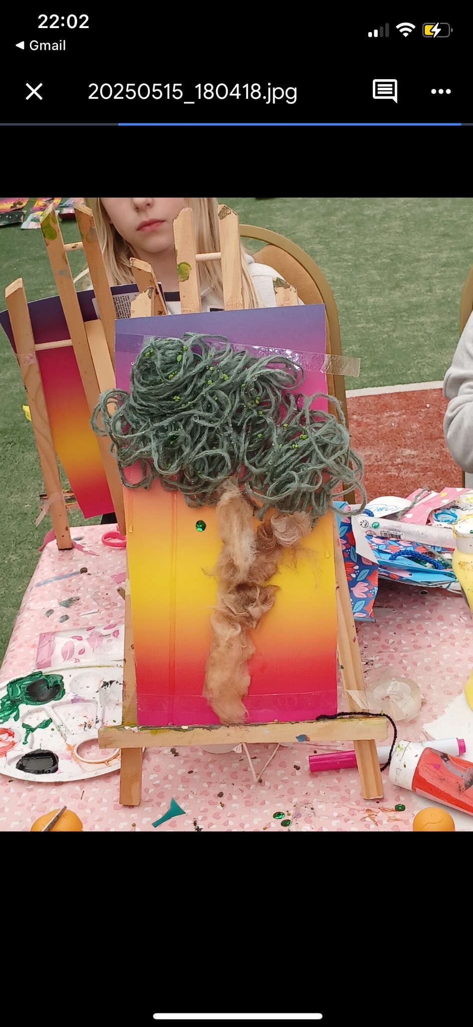 Dziecko tworzy na sztaludze abstrakcyjne drzewo z włóczki i waty na pomalowanym kartonie. Widoczne farby, pędzle i inne materiały plastyczne.