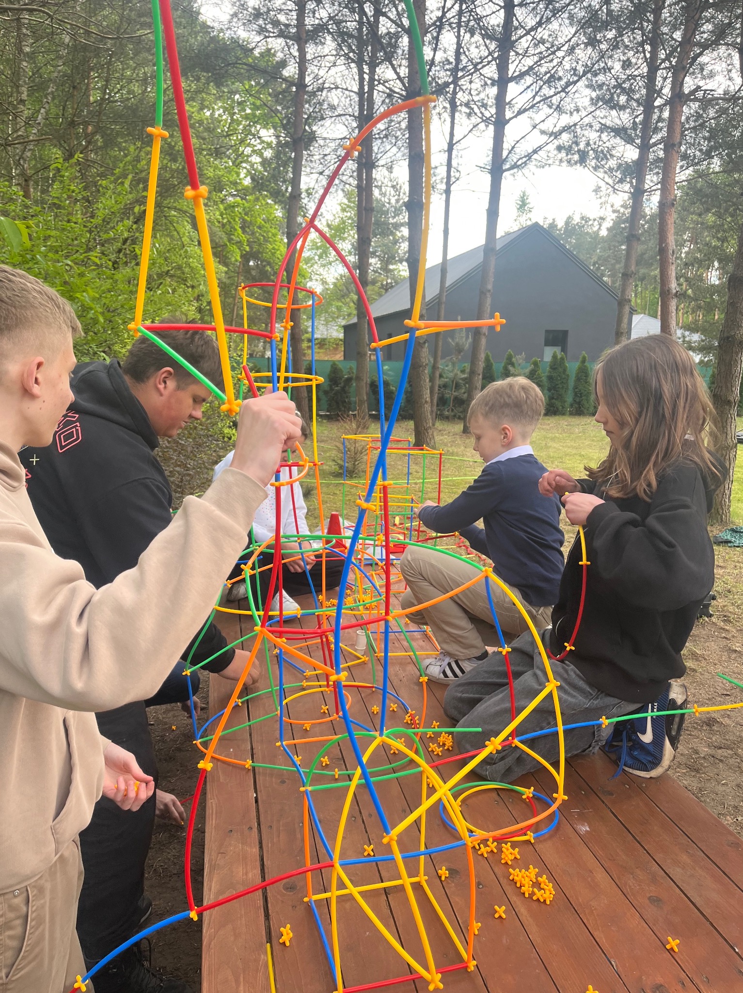 Grupa dzieci i dorosłych buduje kolorową konstrukcję z rurek na drewnianym stole w plenerze. W tle drzewa i dom.