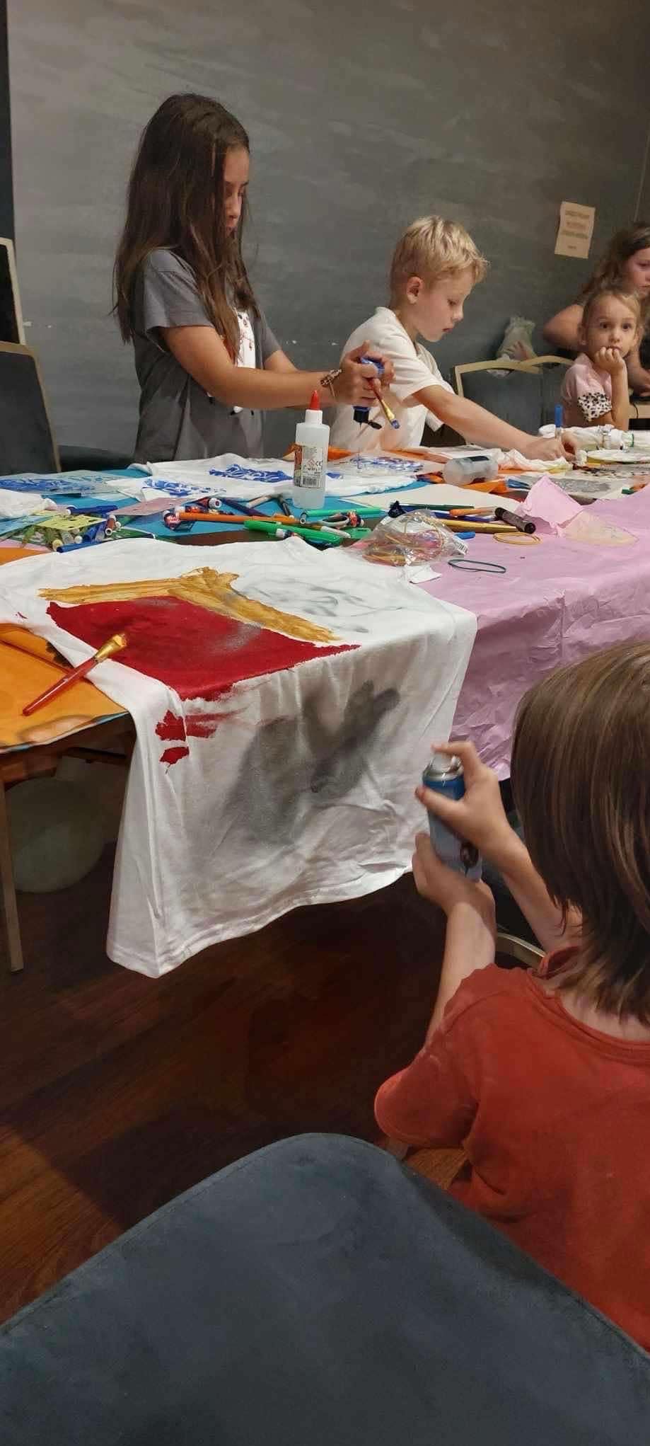 Dzieci na warsztatach artystycznych dekorują białe koszulki farbami. Na stole pędzle, farby i inne akcesoria. Kreatywne zajęcia dla dzieci w pomieszczeniu.