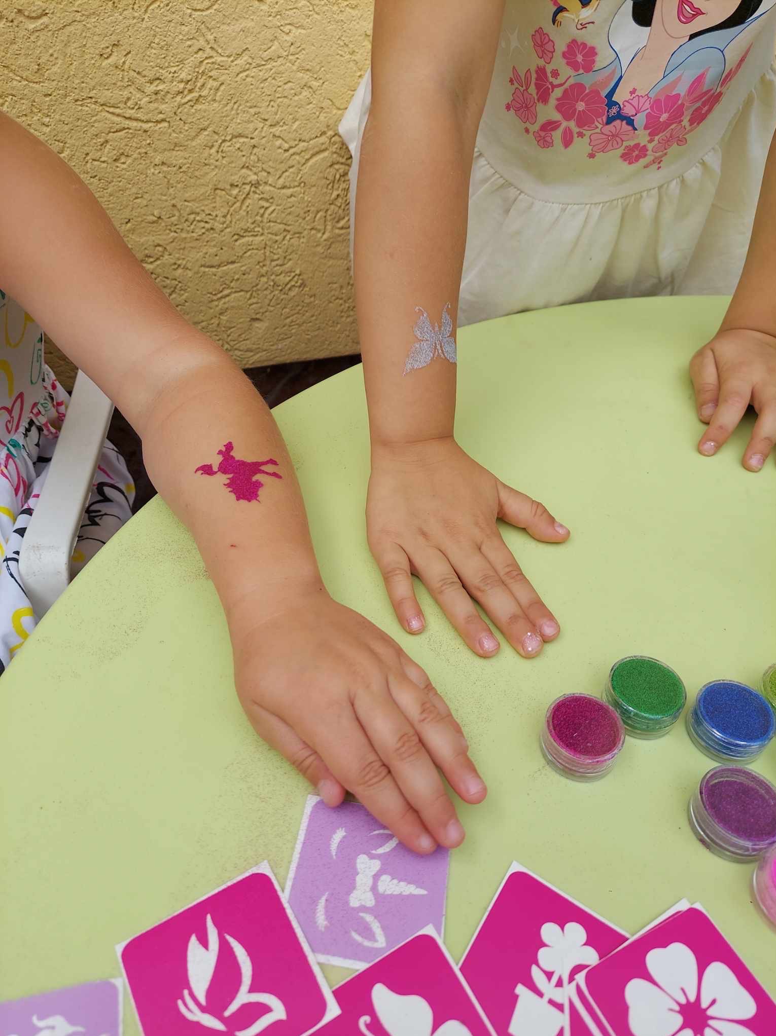 Dzieci z brokatowymi tatuażami na ramionach, siedzące przy zielonym stole z szablonami i brokatem w pojemnikach. Atrakcja dla dzieci.