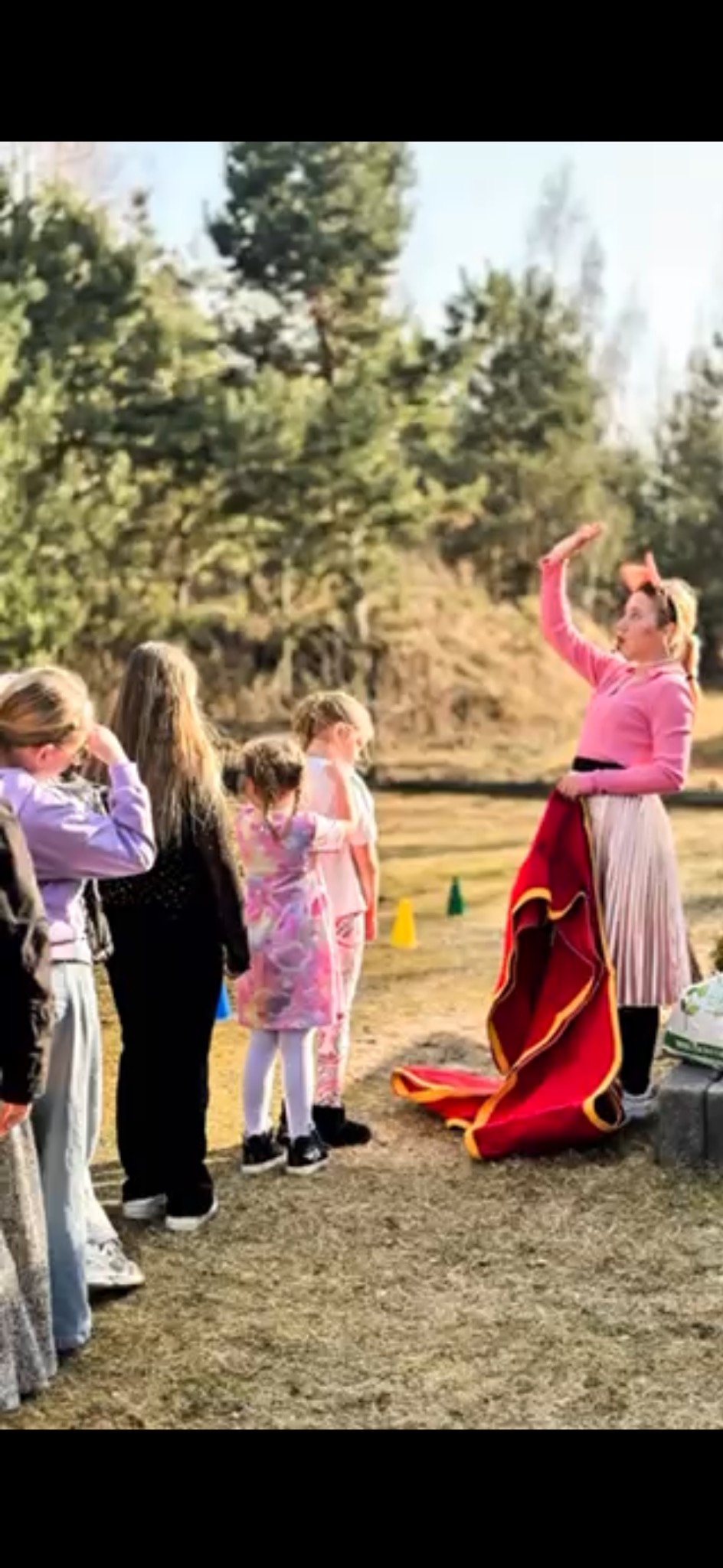 Animator w różowym stroju prezentuje magiczny trik z czerwoną peleryną grupie dzieci na zewnątrz, w tle drzewa i zielona trawa.