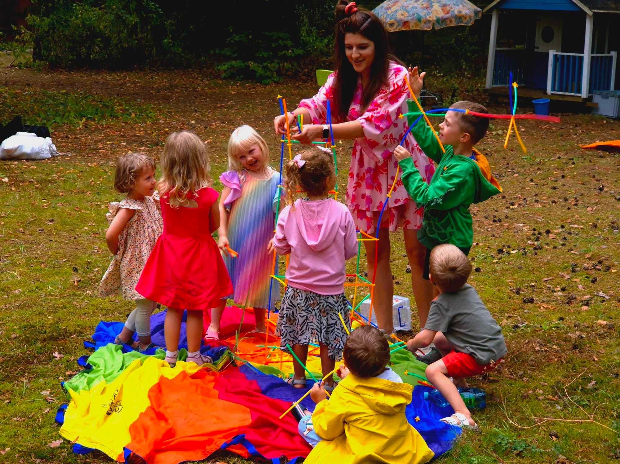 Animatorka w kwiecistej sukience buduje z dziećmi wieżę z kolorowych patyczków na tęczowej chuście w plenerze, z domkiem zabaw w tle.