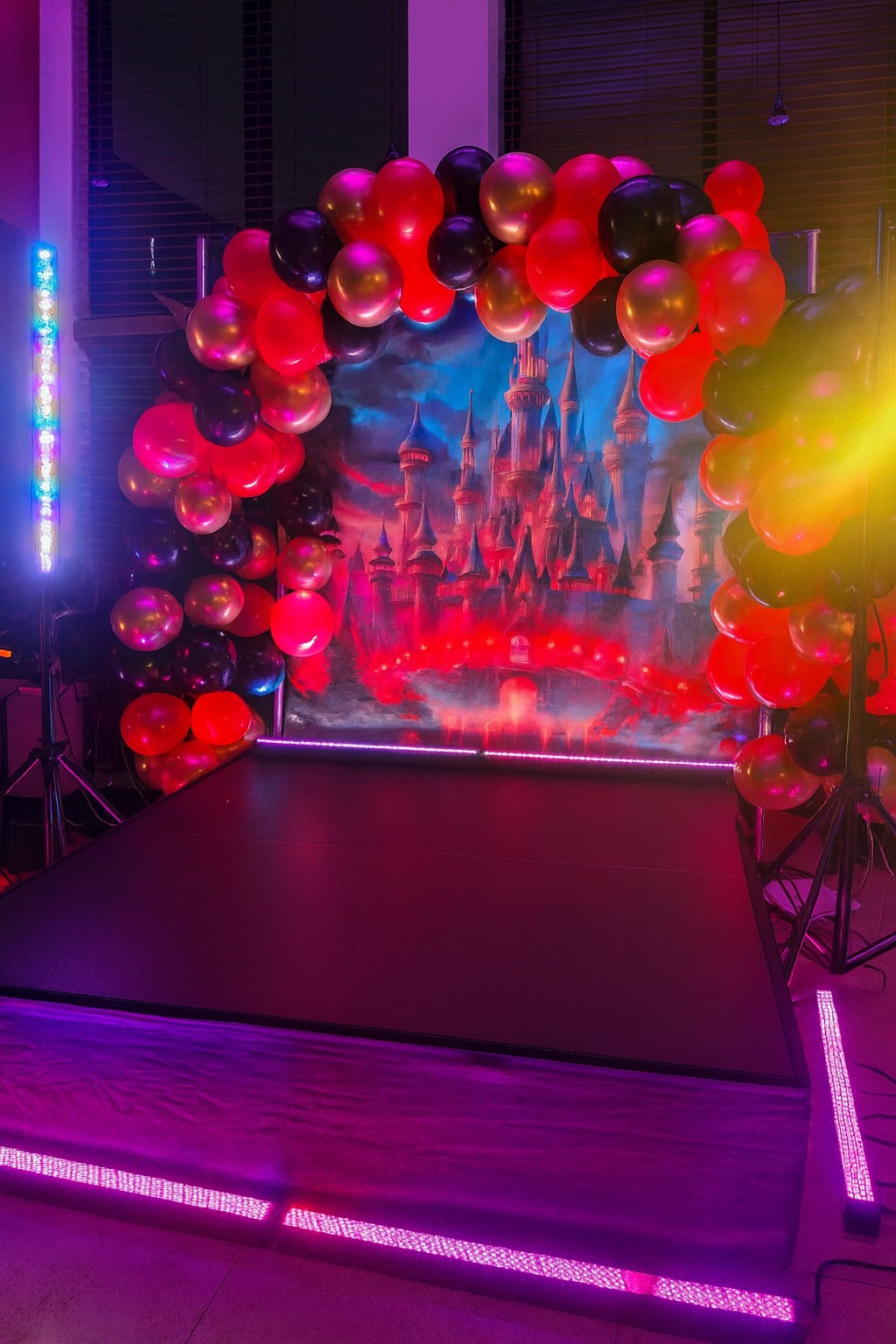 Scena z dekoracją: łuk z balonów (czerwone, złote, czarne), tło z zamkiem, oświetlenie LED w odcieniach różu i fioletu. Gotowa aranżacja na imprezę.