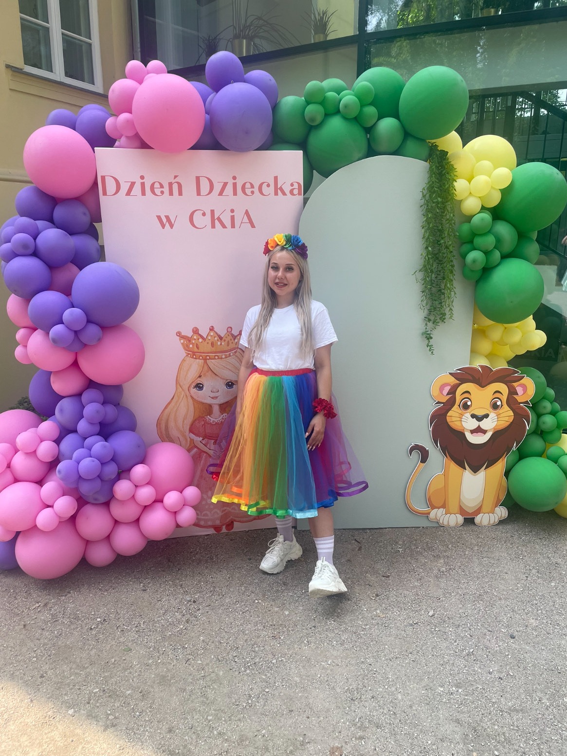 Animator w tęczowej spódnicy pozuje na tle dekoracji balonowych i ścianki z napisem 'Dzień Dziecka w CKIA' oraz rysunkami księżniczki i lwa.