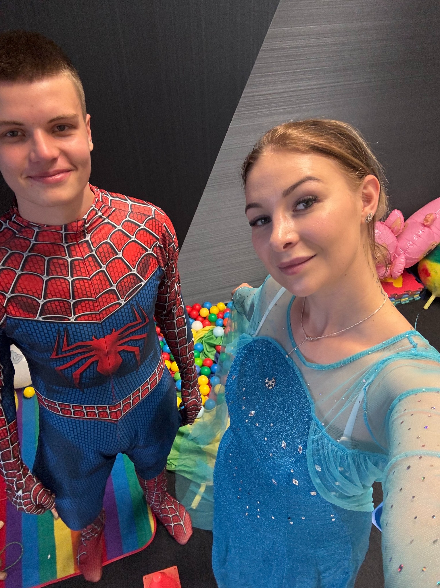Animatorzy w kostiumach: Spider-Man i Elsa, pozują do selfie na tle basenu z kulkami i tęczowego dywanu. Uśmiechnięci, z bliska, w jasnym oświetleniu.