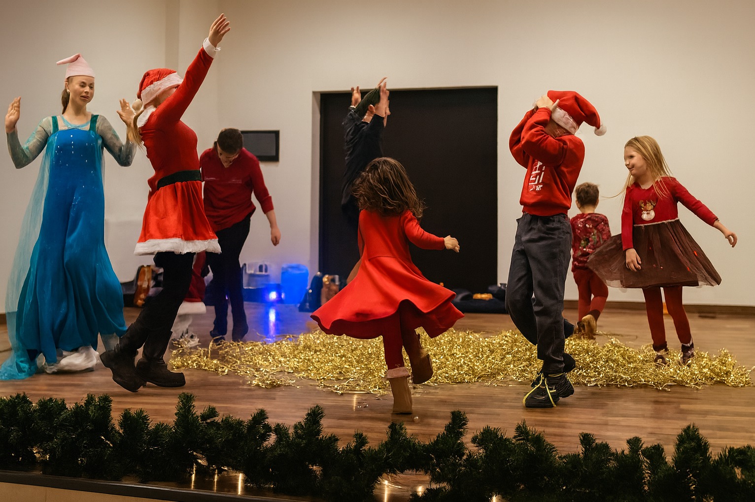 Świąteczny występ dzieci na scenie: dziewczynka w stroju Elsy, dzieci w czerwonych strojach Mikołaja, dekoracje z lampek i girland. Radosna atmosfera.