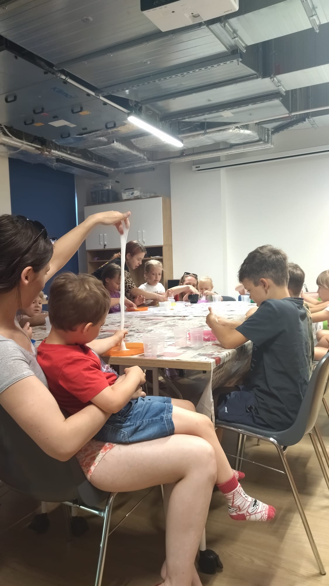 Dzieci przy stole podczas warsztatów, tworzą rozciągliwą masę, widoczna kobieta z dzieckiem na kolanach wskazująca na masę. Sala z widocznymi instalacjami pod sufitem.