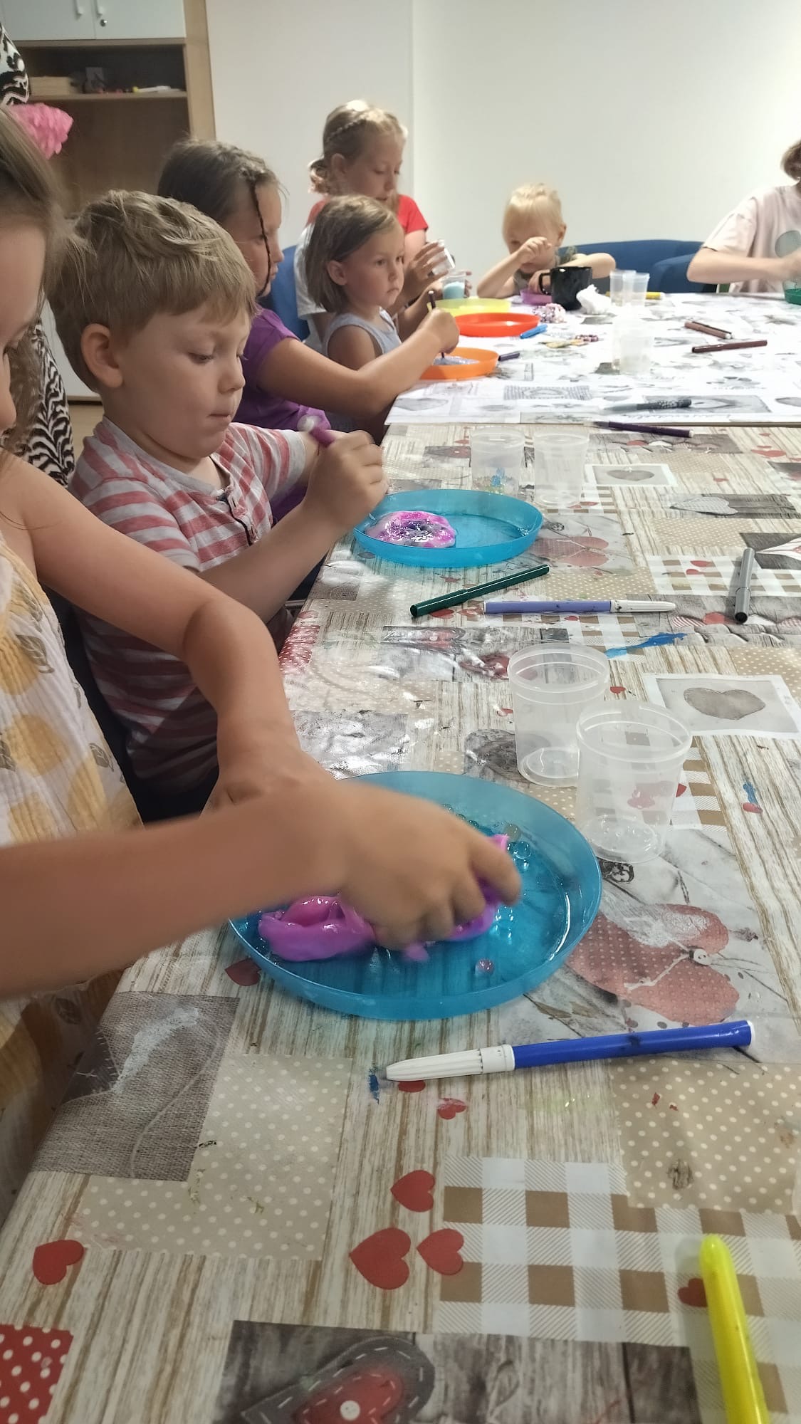 Grupa dzieci siedzi przy stole, tworząc kolorowe masy plastyczne w niebieskich miseczkach. Na stole leżą markery i inne akcesoria plastyczne.