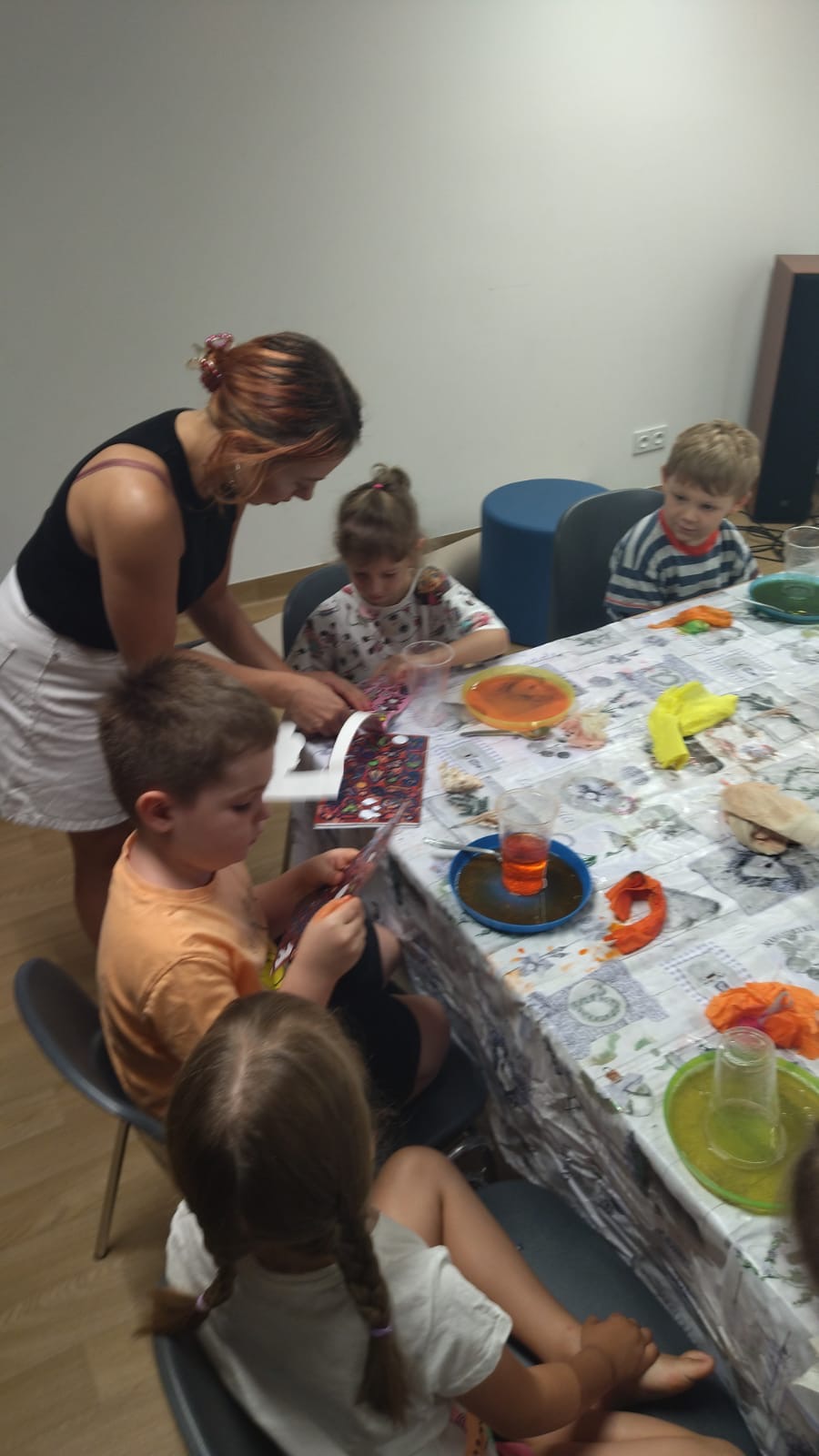 Dzieci i kobieta siedzą przy stole, wykonując prace plastyczne z kolorowych papierów i wycinanek. Stół przykryty ceratą, na nim talerzyki i kubki z płynami.