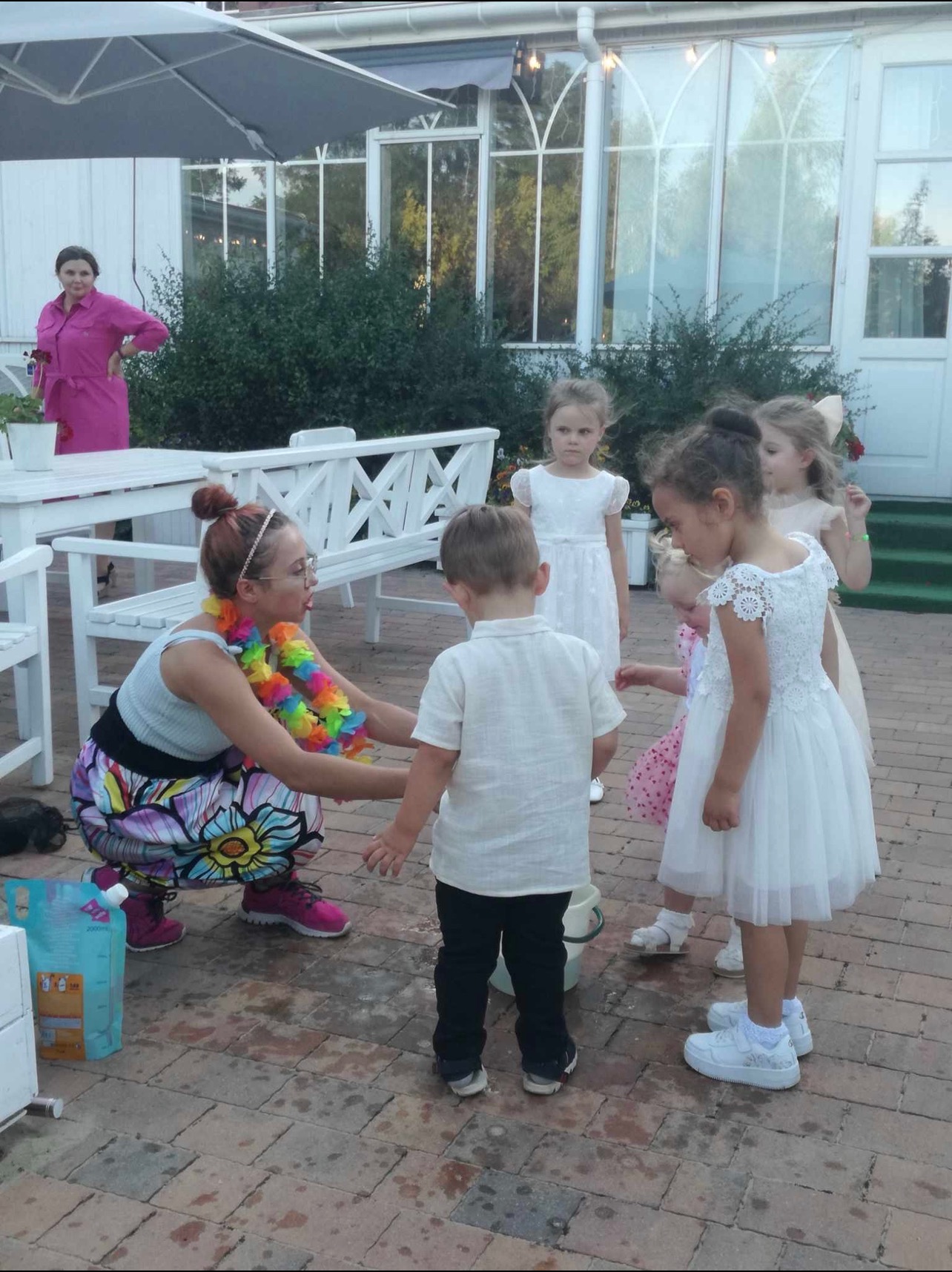 Kobieta w kwiecistej spódnicy i kolorowym naszyjniku rozmawia z grupą dzieci w białych sukienkach na zewnątrz, w tle białe meble ogrodowe i budynek z dużymi oknami.