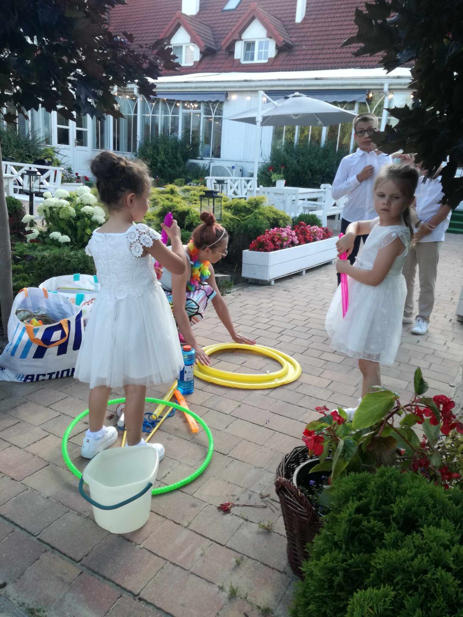 Dwie dziewczynki w białych sukienkach bawią się bańkami mydlanymi i hula-hop na zewnątrz, w tle budynek z białymi elementami architektonicznymi i grupa chłopców w białych koszulach.