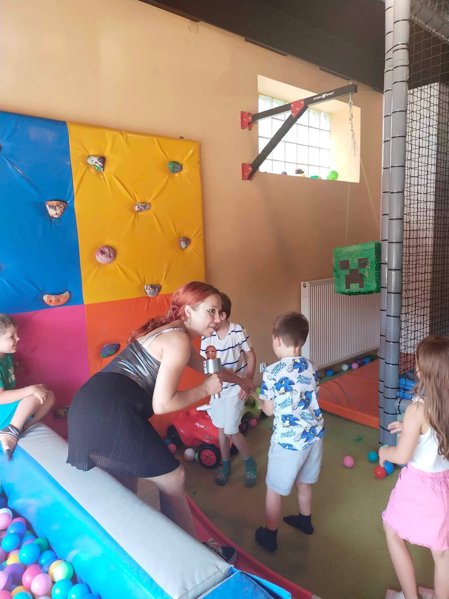 Animator z mikrofonem w srebrnej sukience prowadzi zabawę dla dzieci w sali zabaw z kolorową ścianką wspinaczkową i piłeczkami.