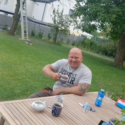 Prace ogrodnicze - Uśmiechnięty mężczyzna z tatuażami, w roboczym ubraniu, siedzi przy drewnianym stole w ogrodzie, trzymając filiżankę. W tle trawnik, drzewa i drabina oparta o drzewo.